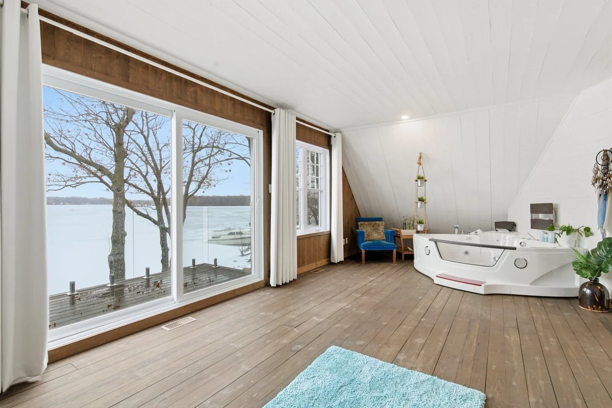 A bright room with large windows overlooking a snowy lake scene, containing a hot tub, blue armchair, potted plants, and wooden flooring.