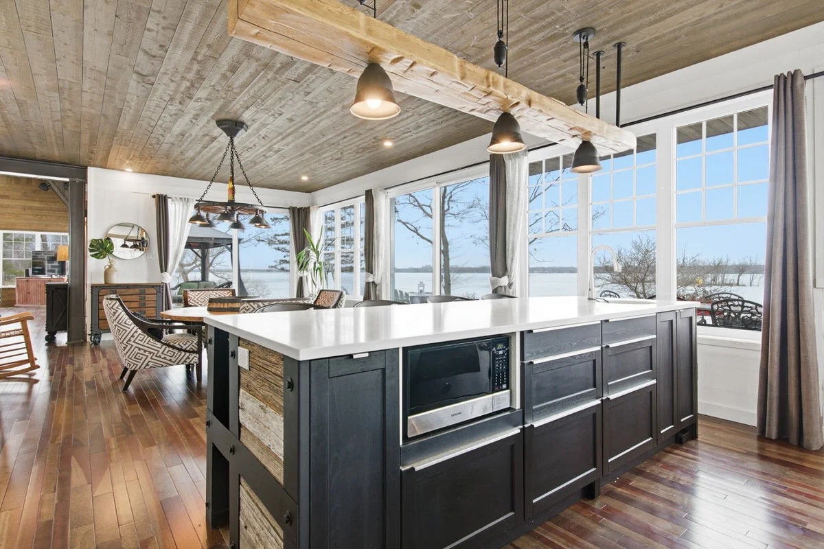 Open concept kitchen and dining area with large windows overlooking a lake. The kitchen features a black island with a built-in microwave, wooden panel accents, and modern black pendant lighting. The dining area has a round table with patterned chair