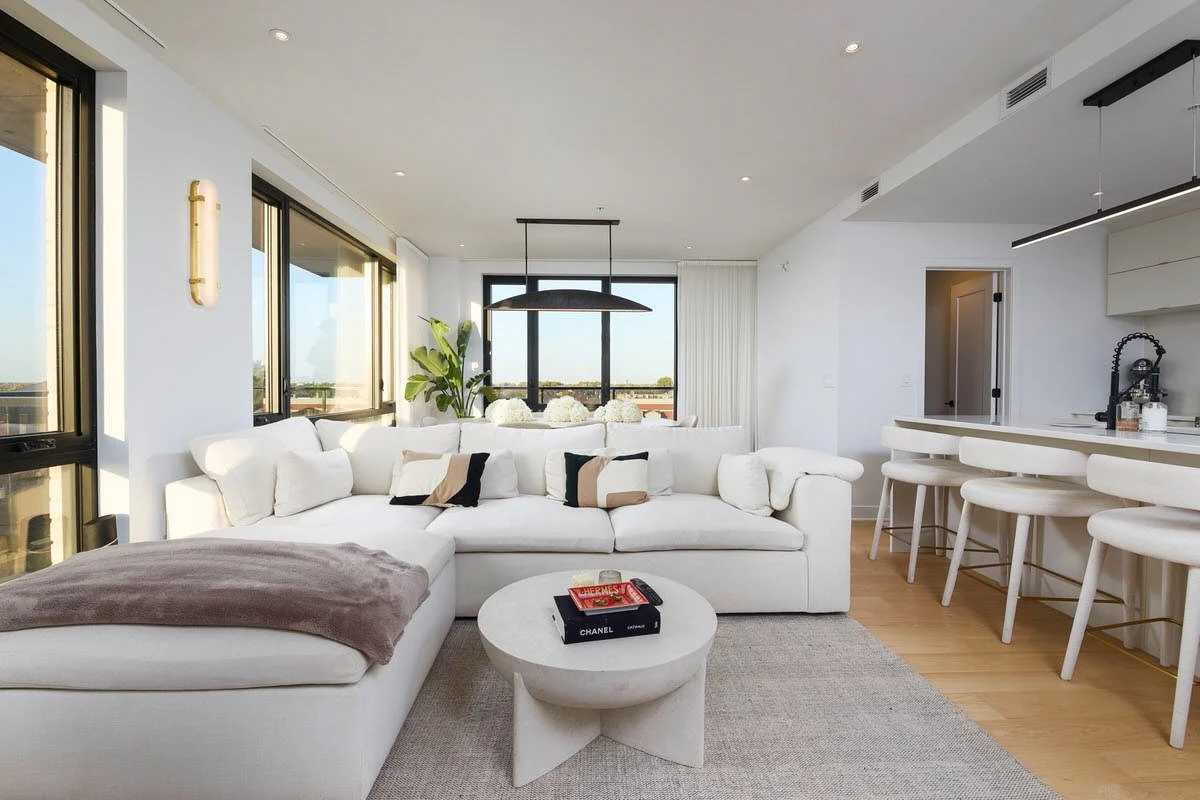Modern living room with white sectional sofa, round coffee table, large windows, hardwood flooring, and bar counter with white stools.
