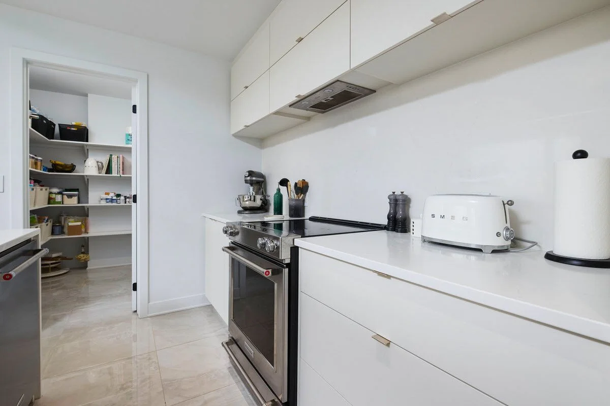 A modern kitchen with white cabinets, a stainless steel stove, and small appliances on the counter, with an open pantry visible in the background.