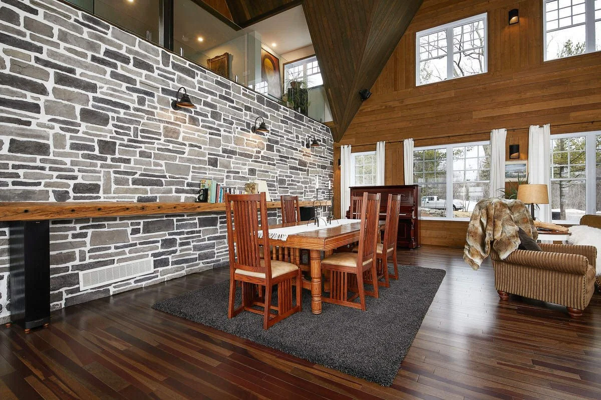 Interior of a modern dining room with a wooden table, six wooden chairs, a stone accent wall with wall-mounted lights, large windows with white curtains, and a vaulted ceiling with wooden paneling.