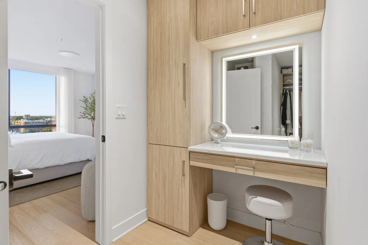 Modern bedroom with a large window, white bedding, and a potted plant, seen through an open door from a white vanity area with a mirror, stool, and storage cabinets.