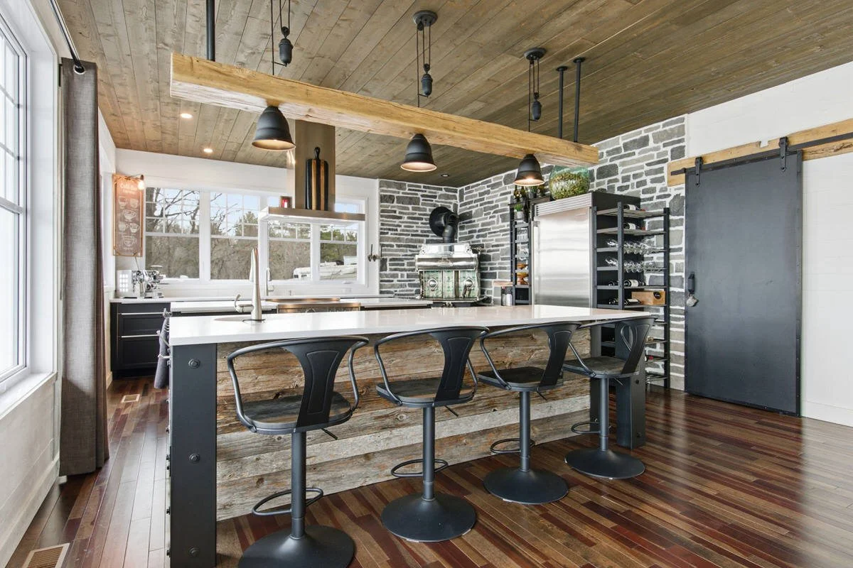 Modern kitchen with a rustic island, black chairs, wood and brick accents, wine rack, and pendant lighting.