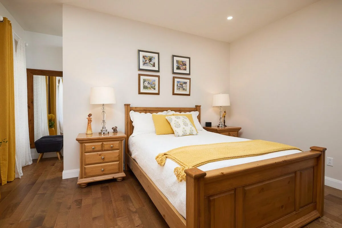 A bedroom with a wooden bed frame, yellow and white pillows, yellow blanket, wooden nightstands with lamps, and framed artwork on the wall.