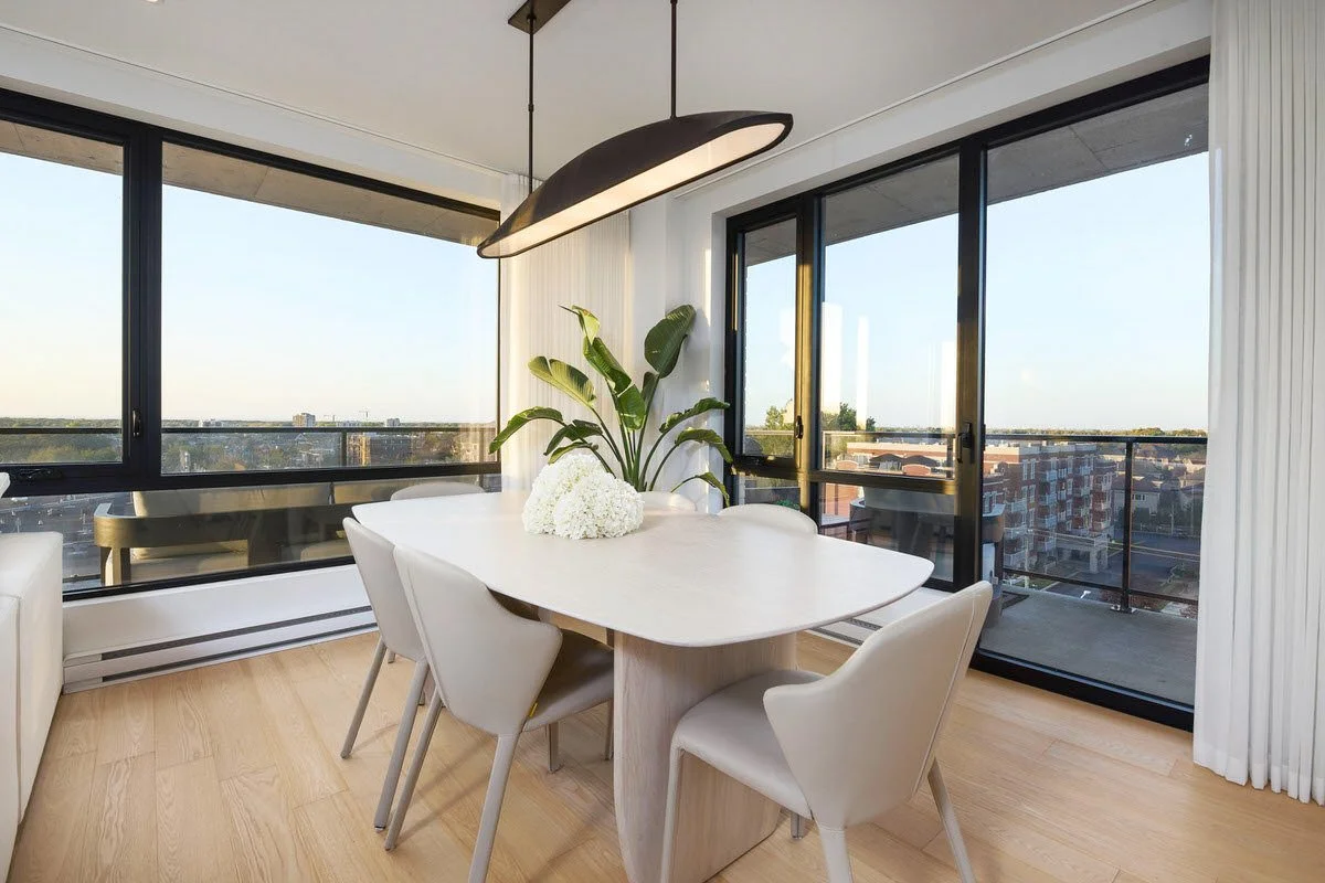 A modern dining room with a white oval dining table, six white chairs, a large green plant, a black pendant light, and large windows with city view.