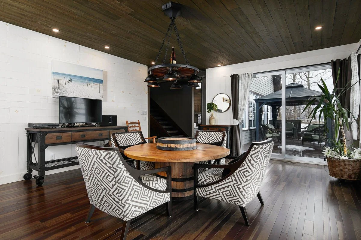 A dining room inside a house with a round wooden table, six patterned chairs, a dark wood ceiling, hardwood floors, a sliding glass door leading outside, and various decorative items.