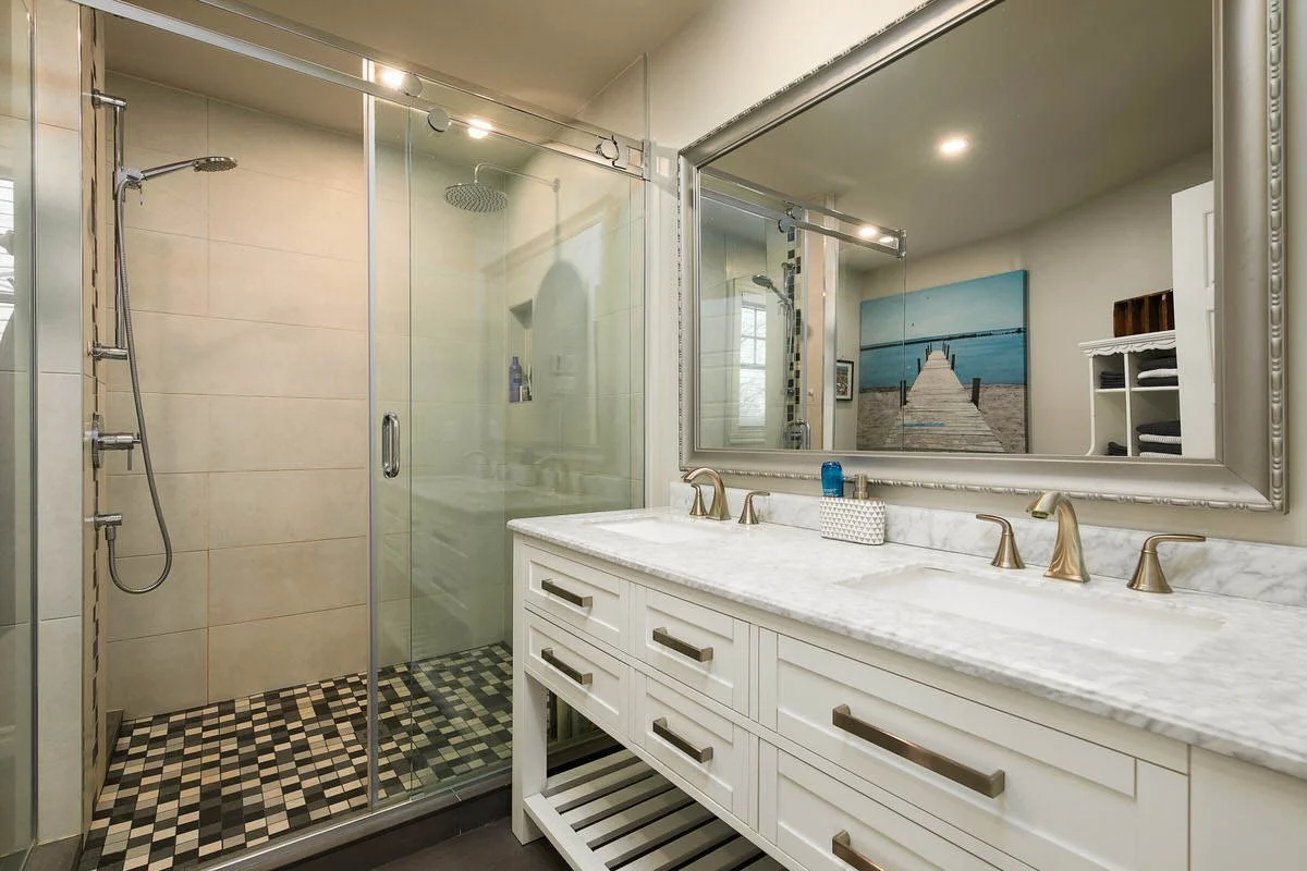 Bathroom with glass shower enclosure and double vanity with marble countertop, large framed mirror, white cabinetry, decorative hardware, and a wall art of a pier leading to the beach.