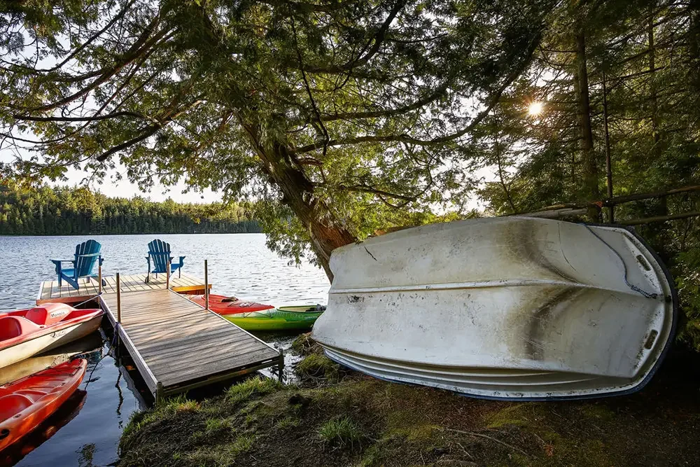 A photo of an overturned boat resting against a tree by a lake with kayaks tied to the dock.
