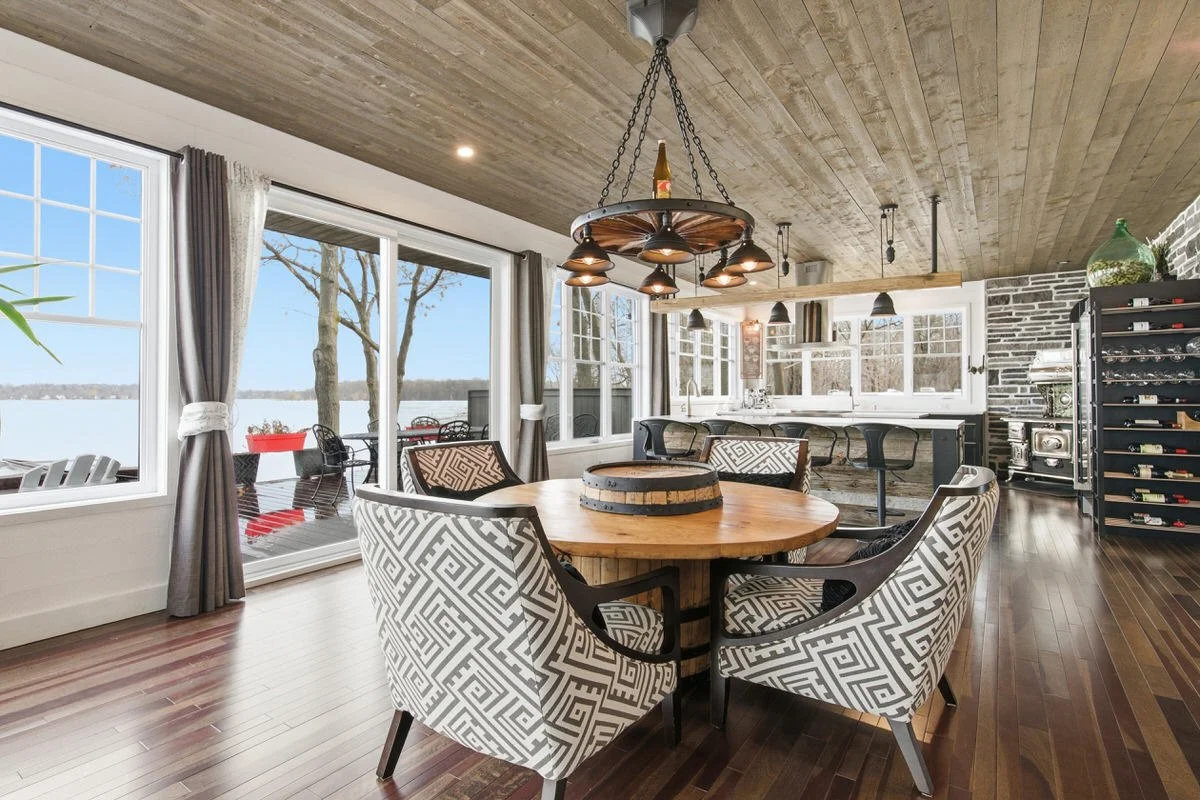 Interior of a dining area with a round wooden table, six patterned chairs, large windows overlooking water and trees, and a kitchen in the background with bar stools, a wine rack, and a stove, featuring wood, brick, and metal decor.