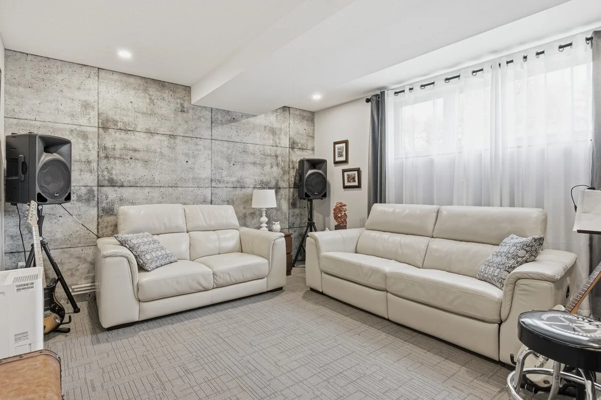 Living room with concrete accent wall, two white leather sofas with patterned pillows, a side table with a white lamp, window with white and gray curtains, and musical equipment including speakers and a guitar.
