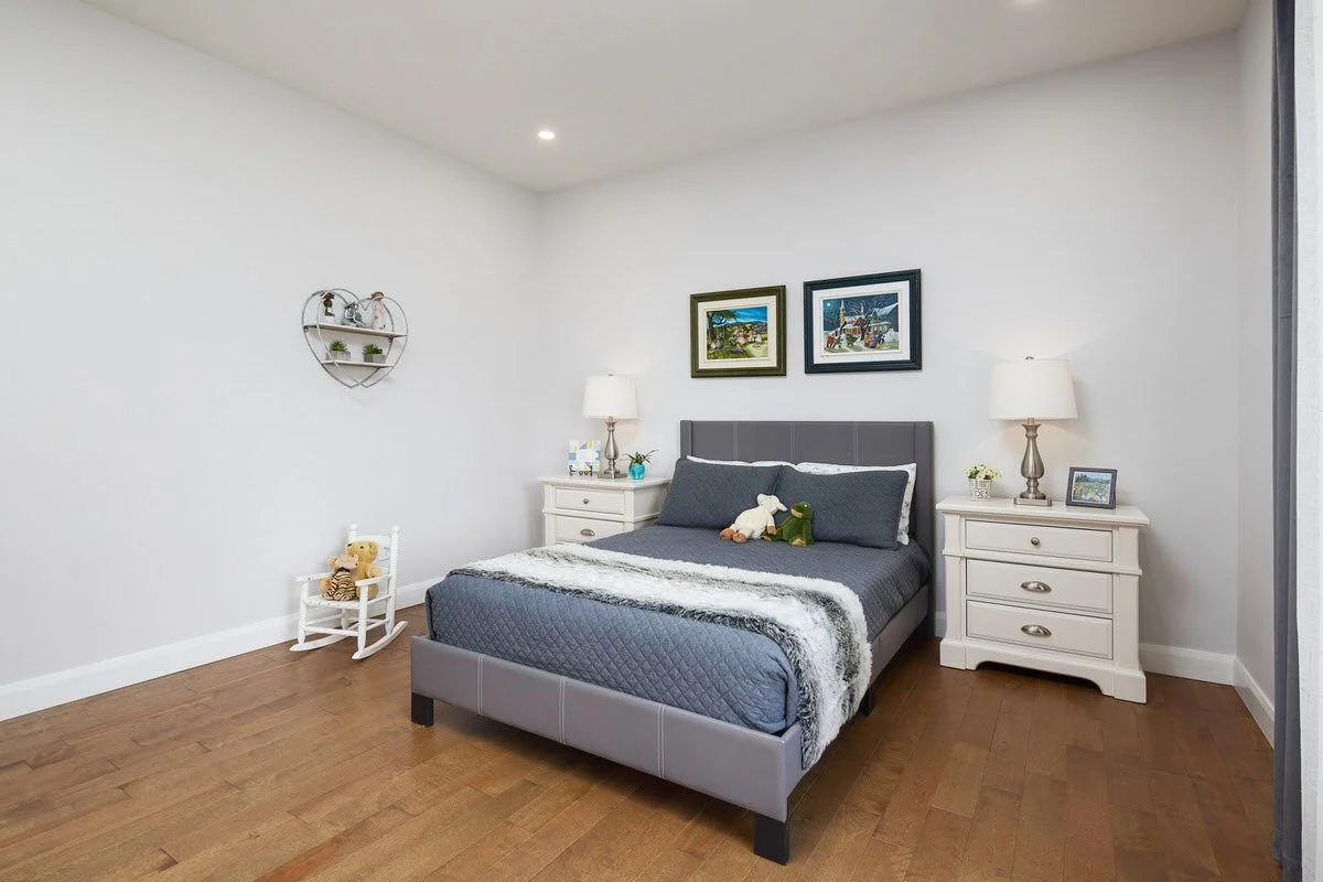 A bedroom with a neatly made bed with dark gray bedding, two plush toys on top, and two white nightstands with lamps. Decorative artwork hangs above the bed, and a small white rocking chair with a teddy bear is in the corner.