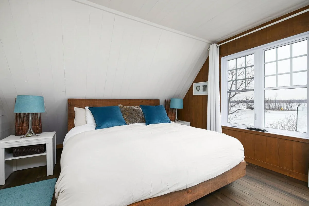 A bedroom with white walls, a wooden bed with white bedding and blue pillows, blue lamps on white nightstands, and a large window showing a winter scene outside.