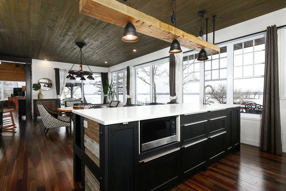Open kitchen and dining area with large windows overlooking a lake, black kitchen island with microwave, wooden ceiling beam, and dark wood floors.