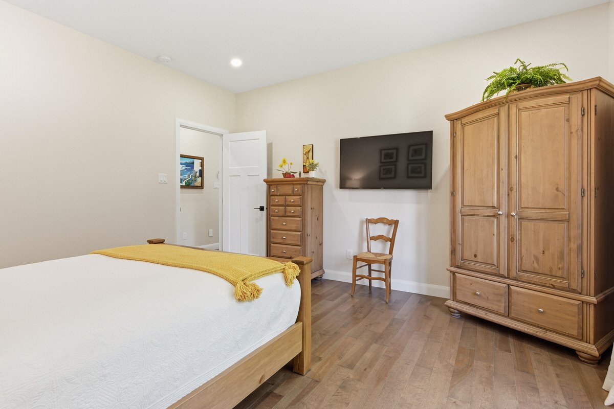 Bedroom with a bed with a yellow throw, wooden wardrobe, chest of drawers, wooden chair, and a wall-mounted TV.