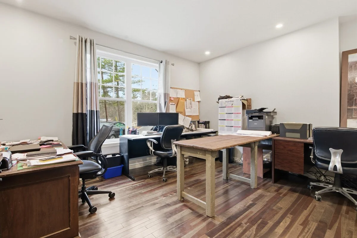 Office room with three desks, office chairs, filing cabinets, and a large window with curtains, some papers and office supplies on the desks.
