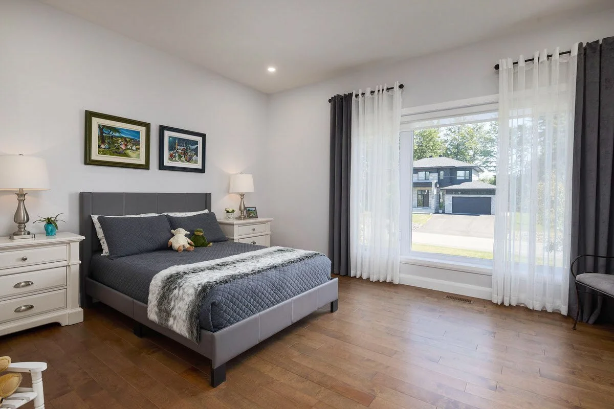 A bedroom with a gray bed, white nightstands, paintings, and large windows with white and dark curtains, showing a house and trees outside.