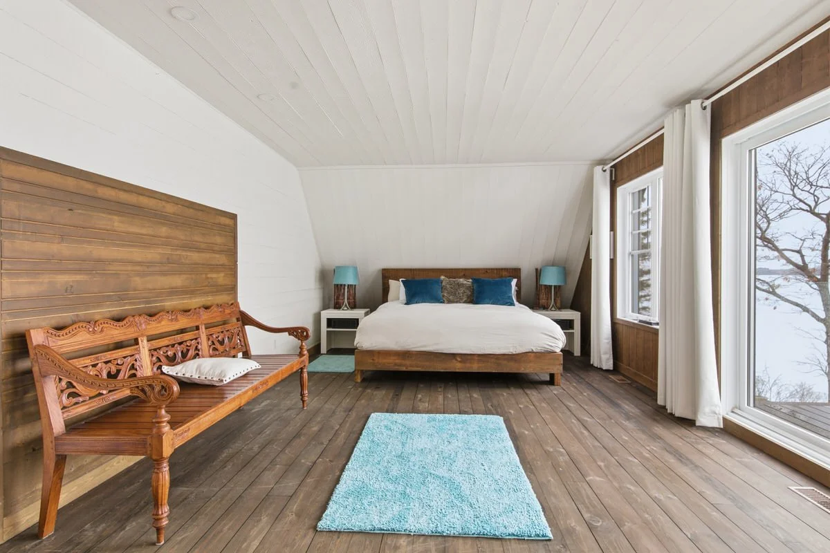 A cozy bedroom with a wooden bed, white bedding, blue and brown accent pillows, two white nightstands with blue lamps, a wooden bench with a cushion, a small dresser, a blue rug, and large windows with white curtains overlooking a tree outside.