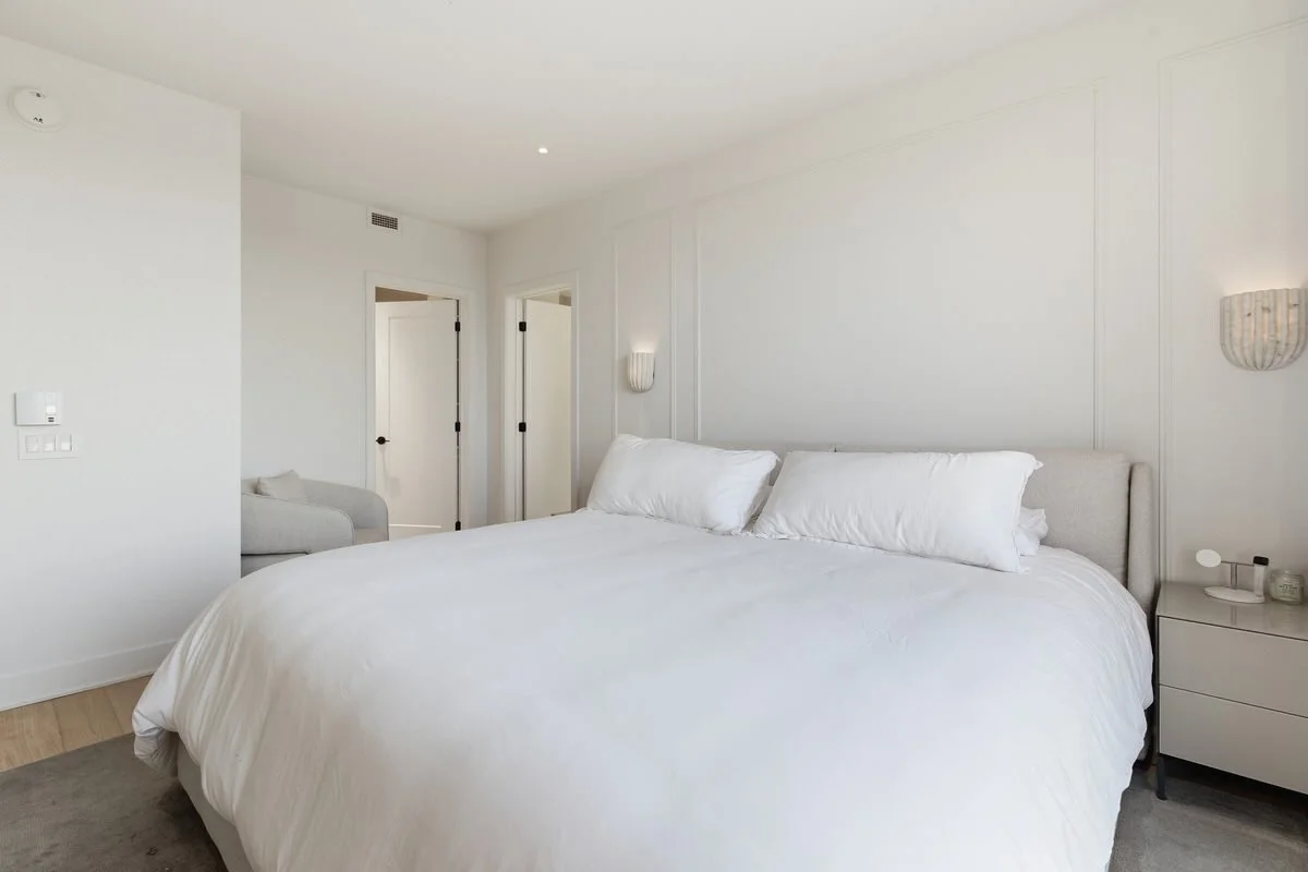 A clean, minimalist bedroom with a large bed, white bedding, and two wall sconces. There is a bedside table with small items and a doorway leading to another room or hallway.