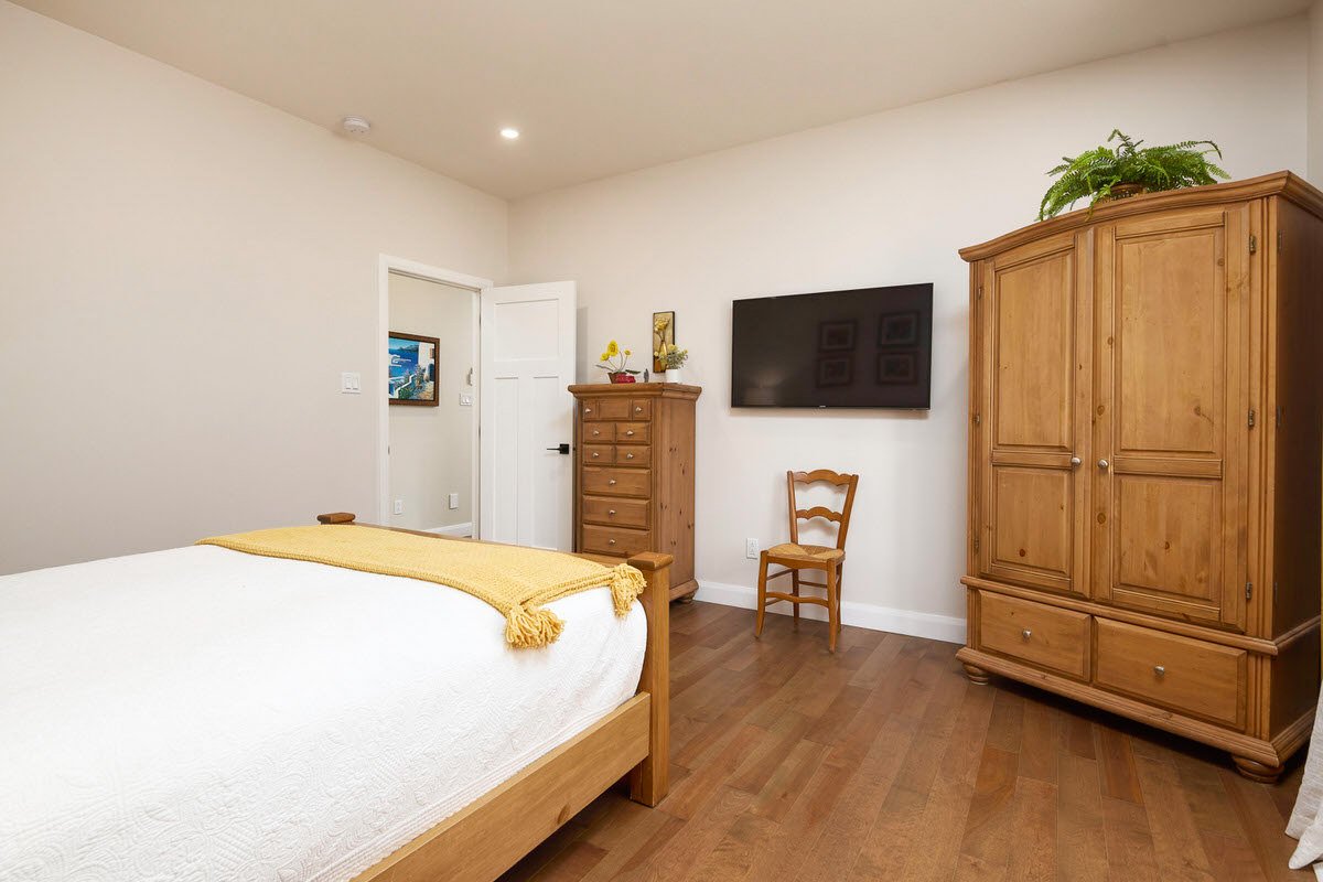 A bedroom with a wooden bed, dresser, and wardrobe, a wall-mounted TV, a chair, and a small bouquet on the dresser.