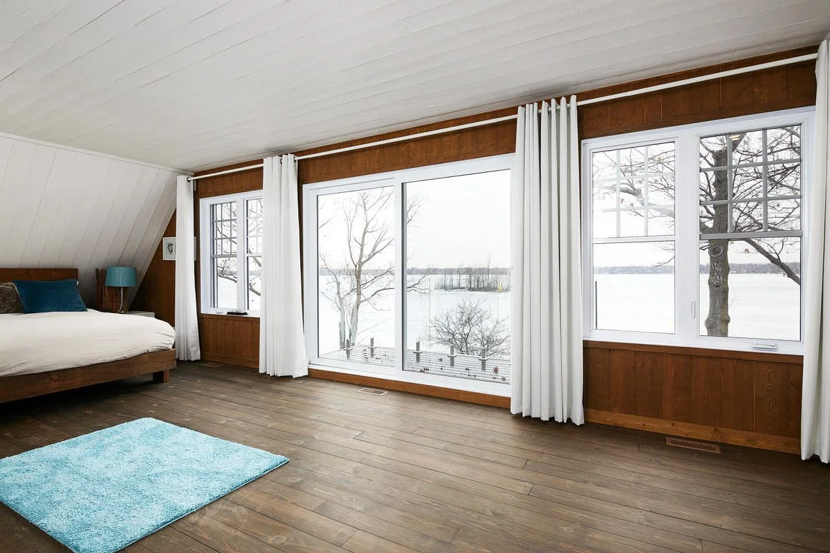 Bedroom with large windows overlooking a snowy landscape and lake, wooden walls, white ceiling, and a blue rug.