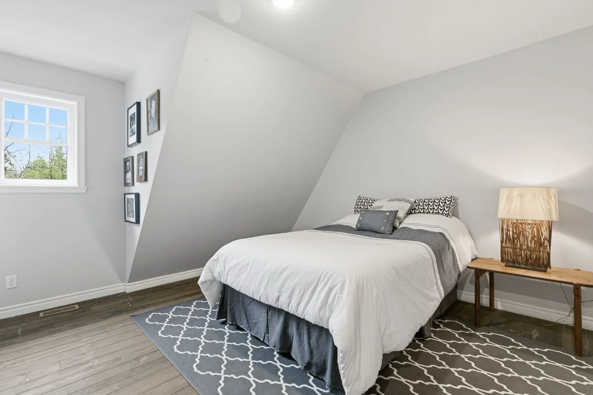A cozy bedroom with white walls and a sloped ceiling, featuring a bed with white and gray bedding, a small wooden side table with a wicker lampshade, and a patterned gray and white rug. A window shows a sunny day outside, and picture frames are hangi