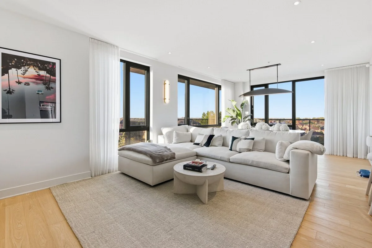 Bright living room with large white sectional sofa, cream rug, coffee table with books, and floor-to-ceiling windows with white drapes, overlooking a cityscape.