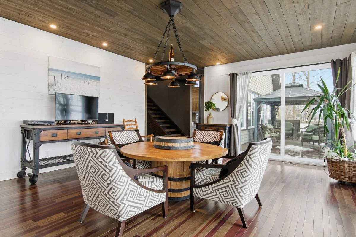 A dining room with a round wooden table, four patterned chairs, a large window with curtains, a sliding door leading outside, a wooden ceiling, and a chandelier hanging above the table.