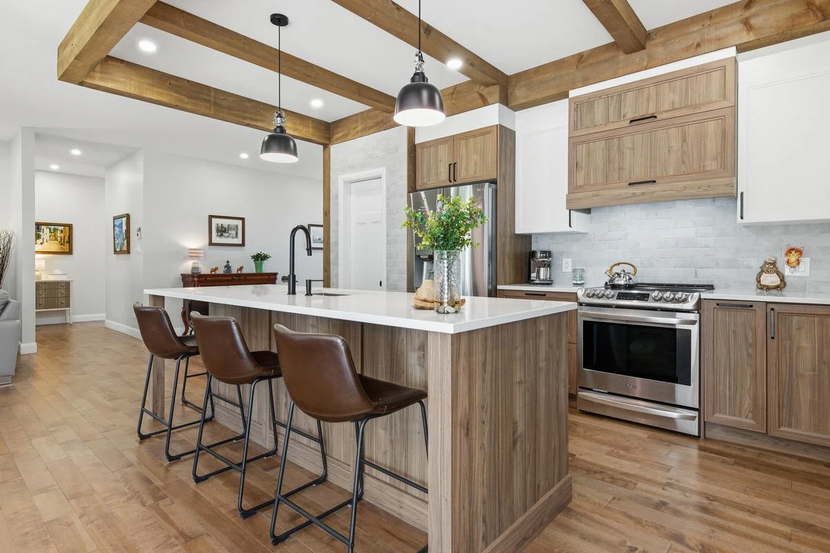 Modern kitchen with wooden ceiling beams, a white island counter with a vase of green plants, brown leather chairs, stainless steel appliances, white and wooden cabinetry, and hardwood floors.