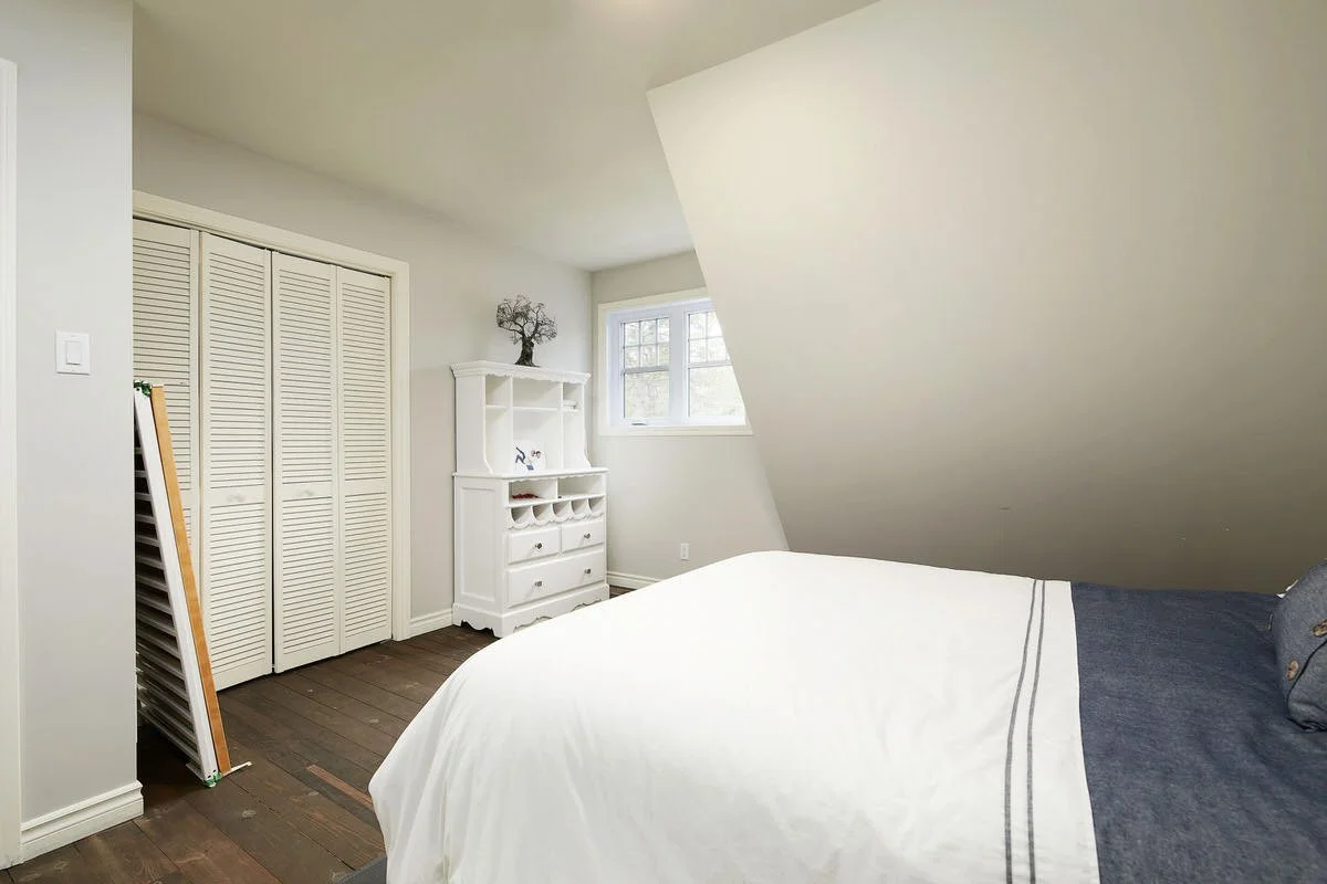 A bedroom with a bed covered in white and dark blue bedding, a white dresser, a small window, and a closet with louvered doors.