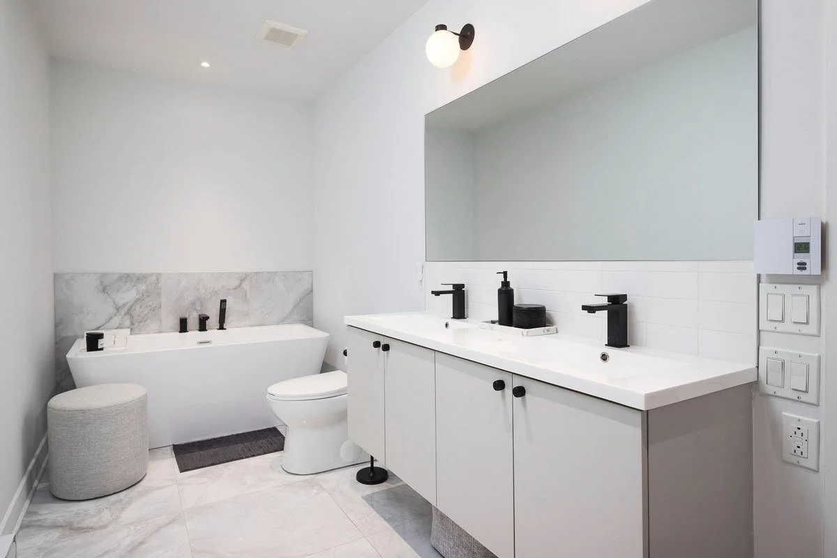 Modern white bathroom with a large vanity, two black faucets, a large mirror, a bathtub with black fixtures, and gray accent wall behind the tub.