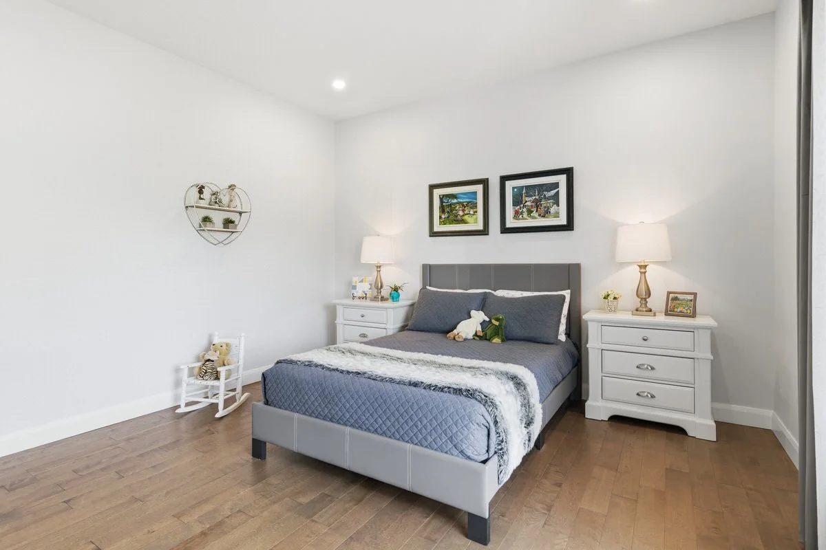 A modern bedroom with a gray upholstered bed, white nightstands on each side, and décor including framed pictures, lamps, and small decorative items. There are plush toys on the bed and a small stuffed animal on a chair at the side.