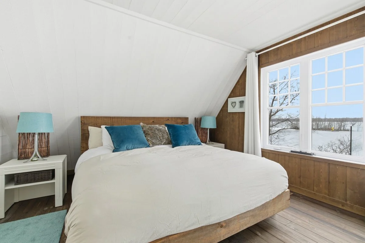 A cozy bedroom with a large white bed, blue pillows, and a rustic wooden headboard, situated near a window with scenic winter view.