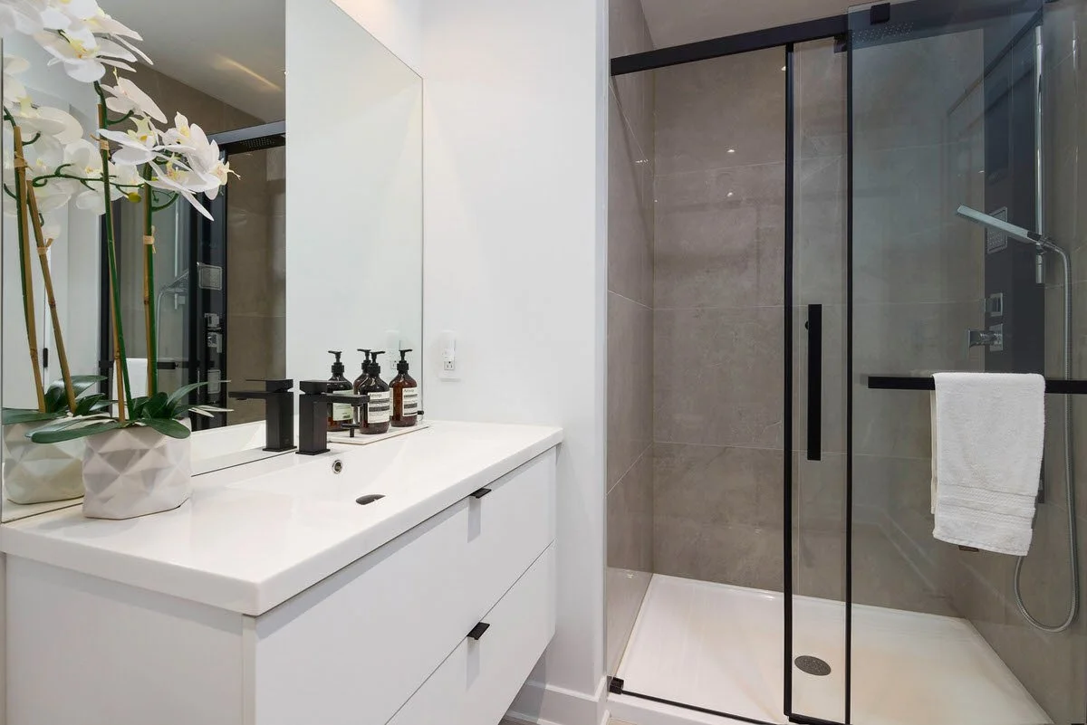 Modern bathroom with a white vanity, black faucet, potted orchid, and a glass shower with a towel hanging on the door.