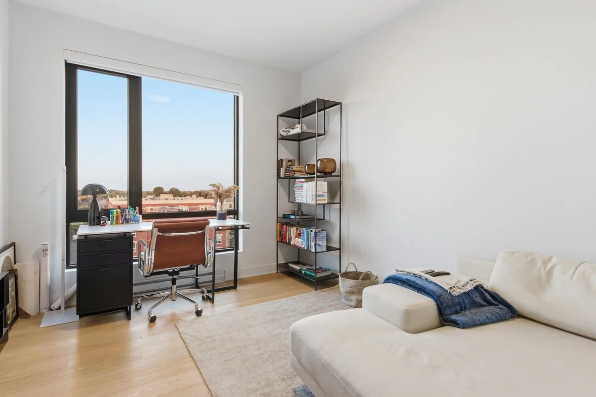 A bright, minimalist home office and living room with a large window, desk with office supplies, an office chair, a tall black bookshelf filled with books and decorative items, a white sofa with a blue blanket, and a beige rug on light wood flooring.