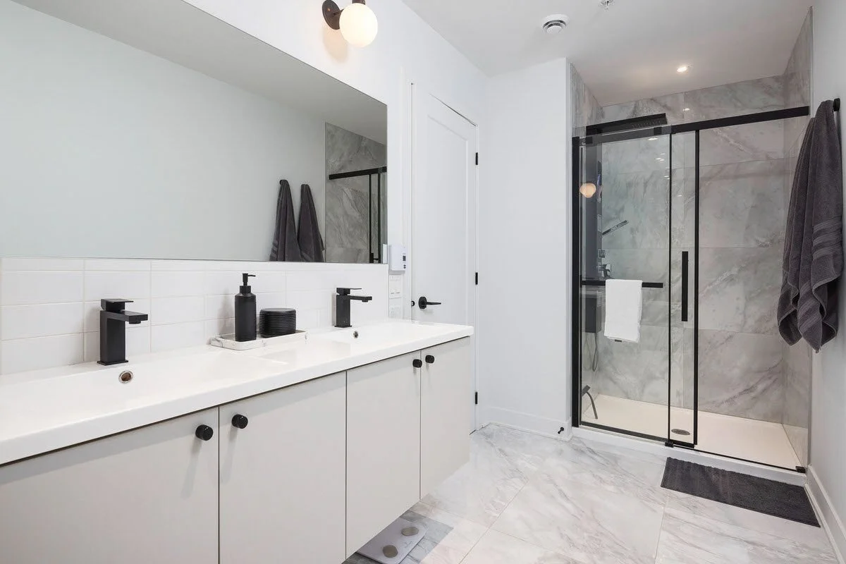 Modern bathroom with double sink vanity, black fixtures, large mirror, and walk-in shower with glass doors and marble walls.