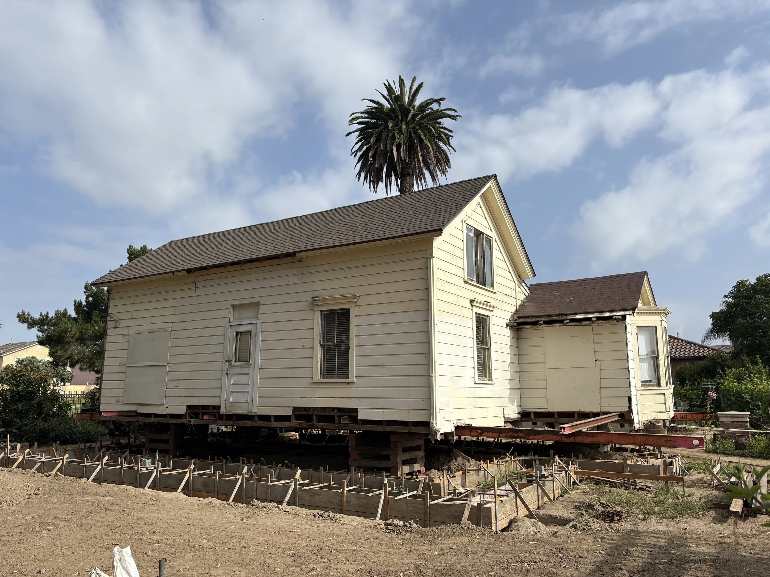 A house is raised above the ground on wooden and concrete supports in a yard during renovation or relocation, with visible weathered white siding and a gray roof, under a blue sky with some clouds and a tall palm tree behind it.