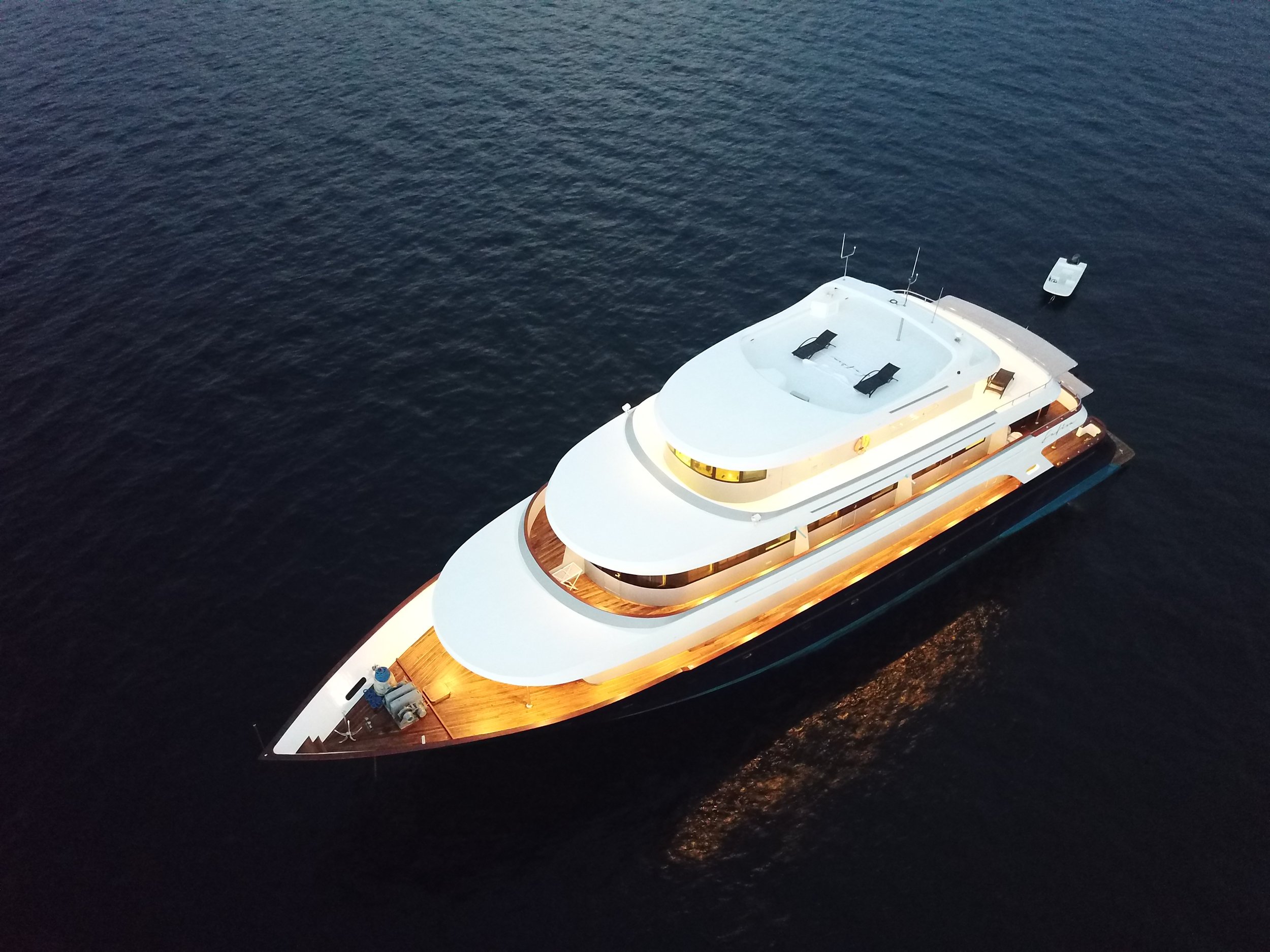 Aerial view of a luxury white yacht with multiple decks, floating on dark water with a small boat nearby.