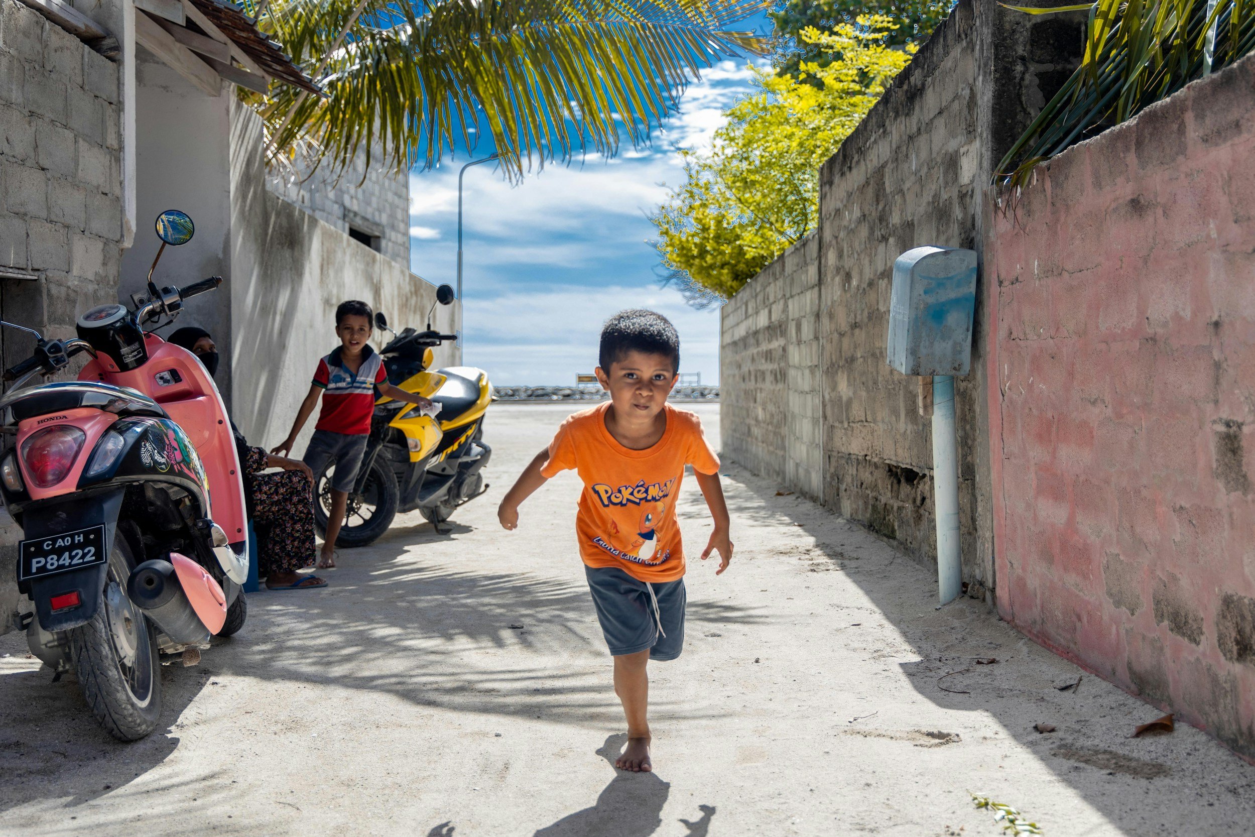 A young boy in an orange Pokémon T-shirt runs towards the camera on a sandy street. Two other children are in the background next to motorcycles, with one of them standing and smiling. The scene is in a sunny area with a pink concrete wall on one side, a white building on the other, and palm trees overhead.