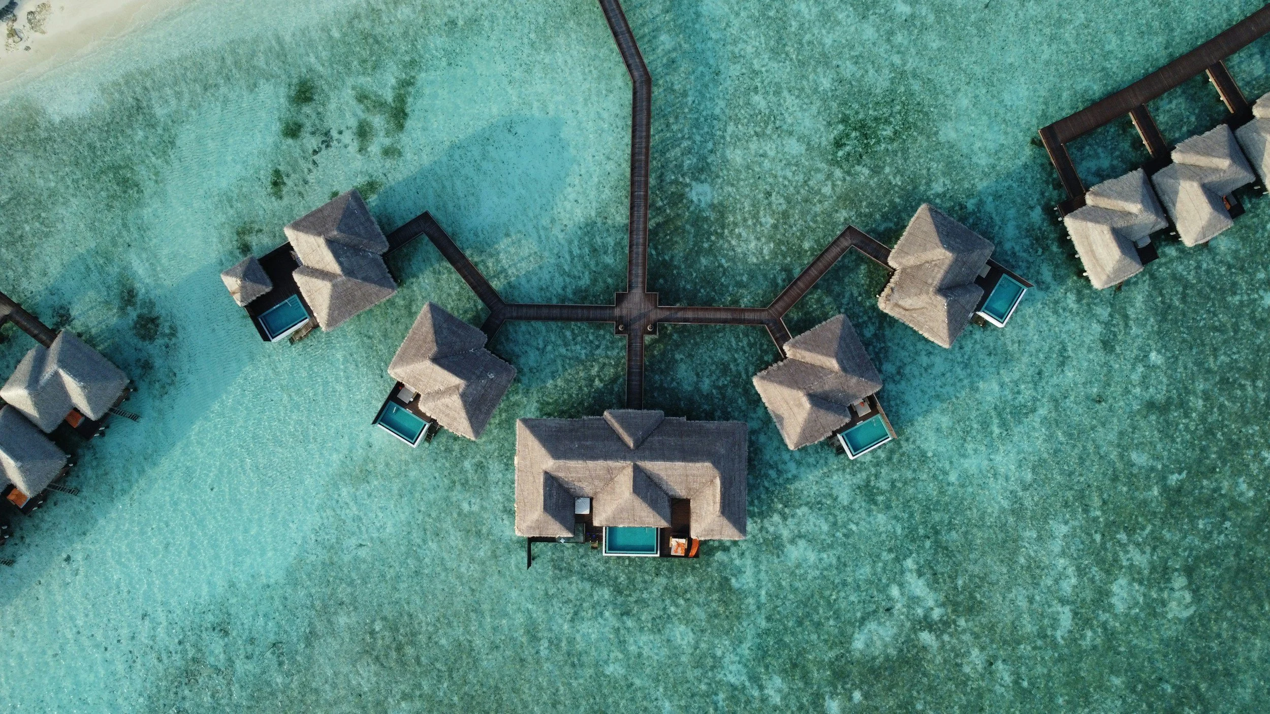 Aerial view of overwater bungalows with thatched roofs connected by a wooden walkway, situated over clear turquoise ocean water.