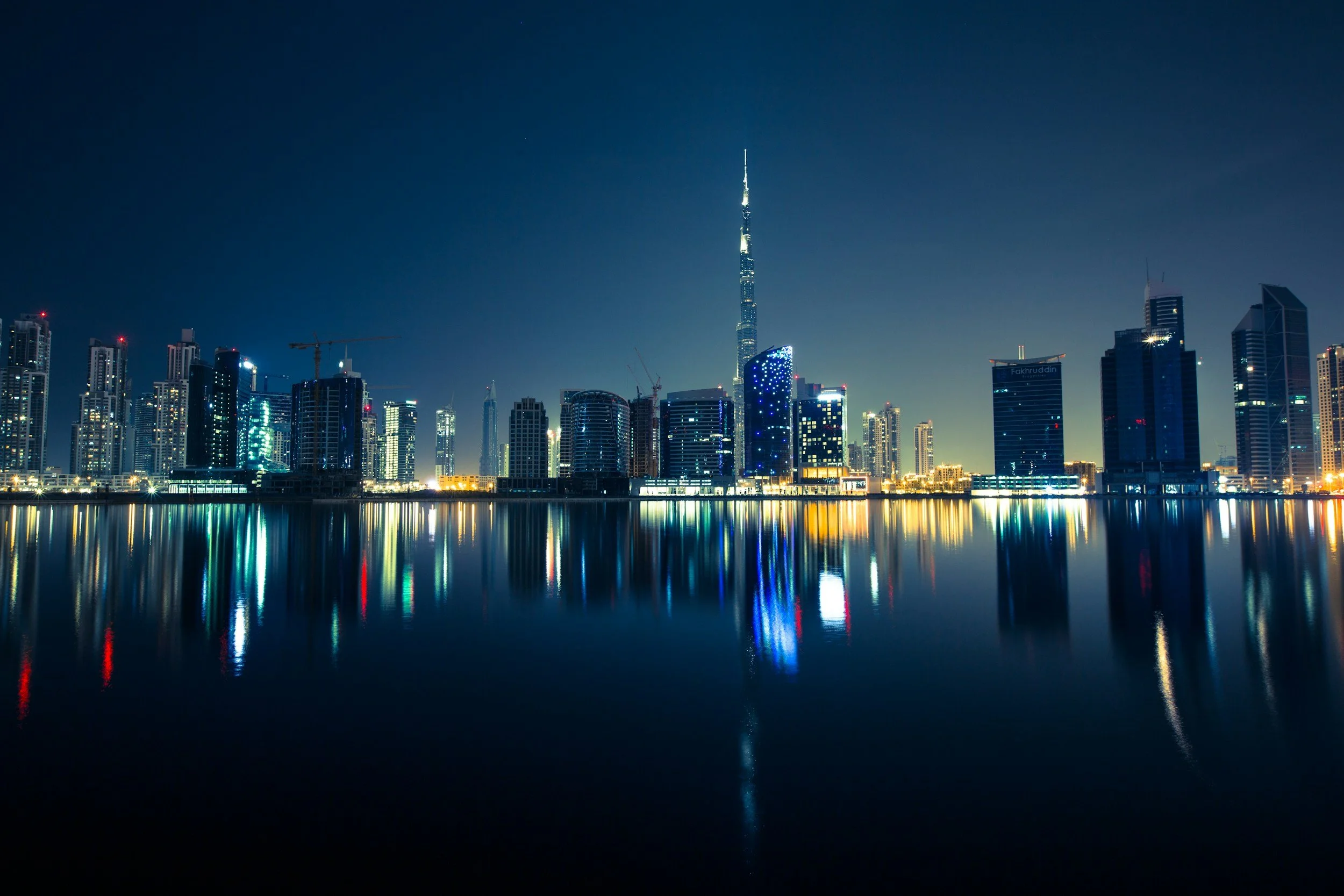 Nighttime city skyline with illuminated skyscrapers reflecting on calm water, featuring the tall Burj Khalifa in Dubai.