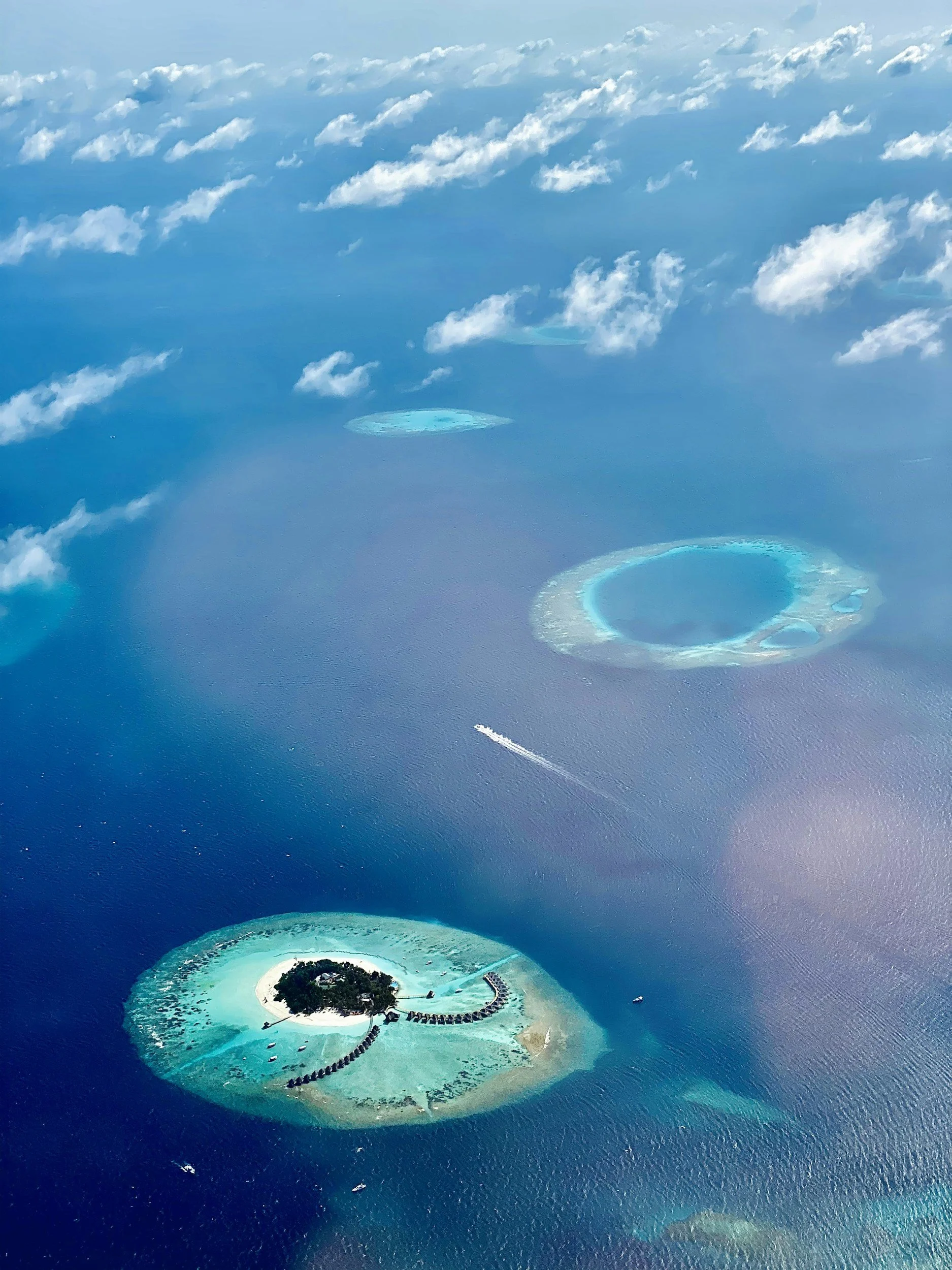 Aerial view of a tropical island with overwater bungalows, surrounded by turquoise water and coral reefs, with smaller atoll islands in the distance.