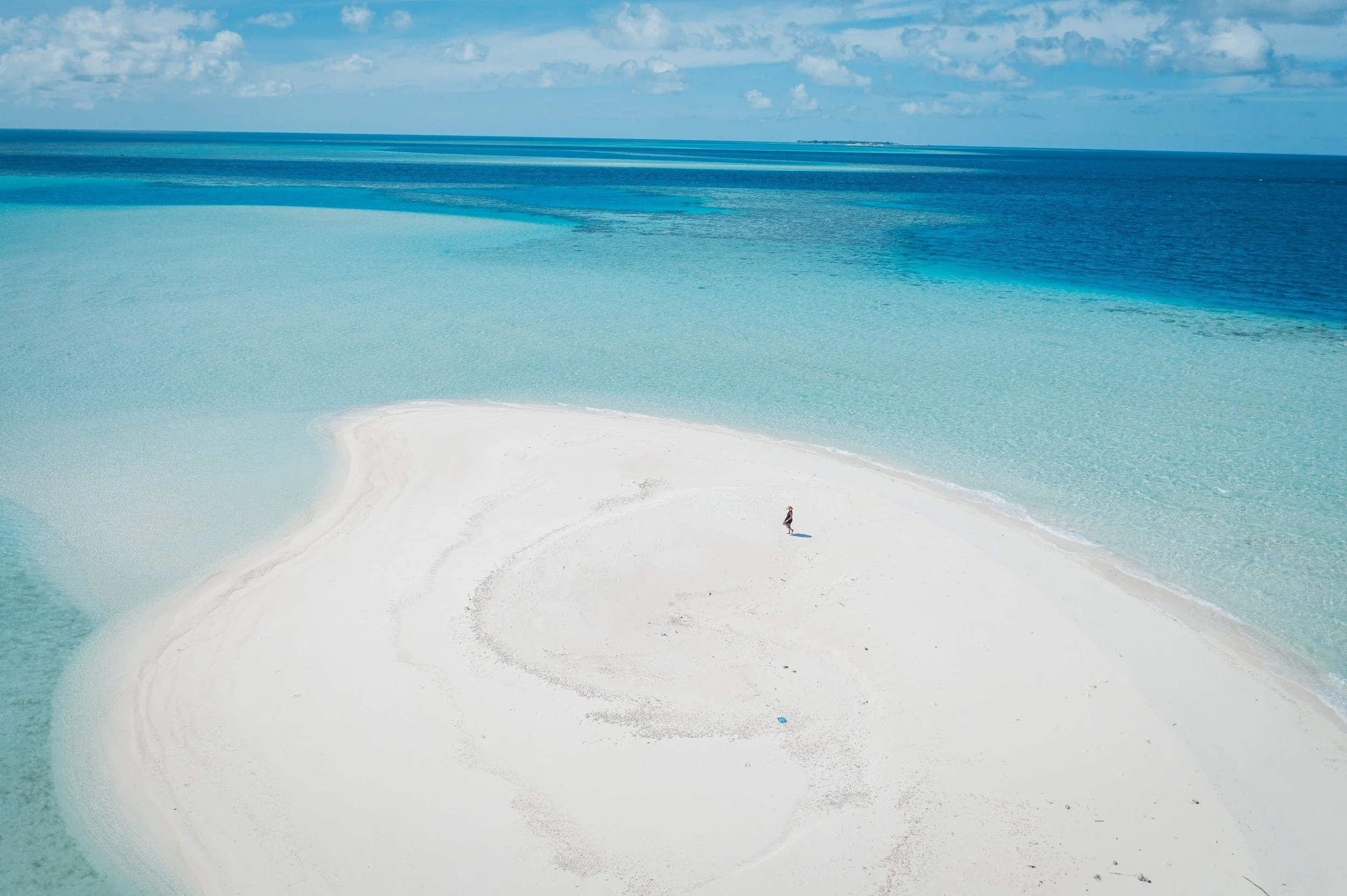 A person walking on a white sandbar surrounded by turquoise blue waters near the ocean, under a partly cloudy sky.