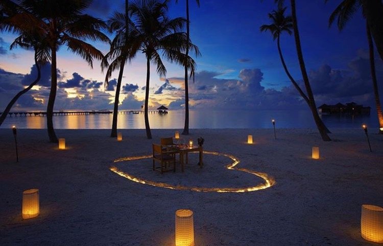 Beach scene at sunset with palm trees, a table set for dinner, and tiki torches along the shore with a pier in the background
