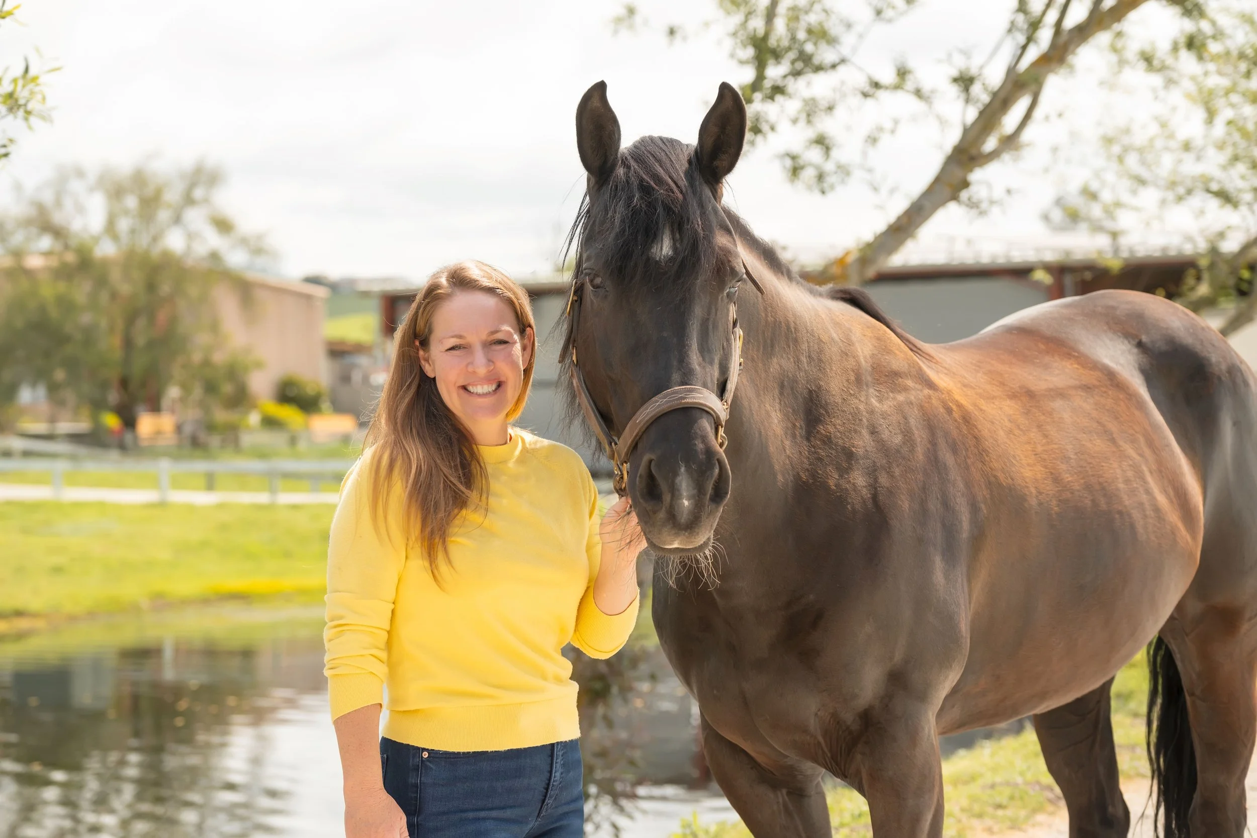Maggie K. Clancy, owner of Strides Riding Academy, standing beside a horse in a riding arena, smiling and sharing a calm connection with the horse.