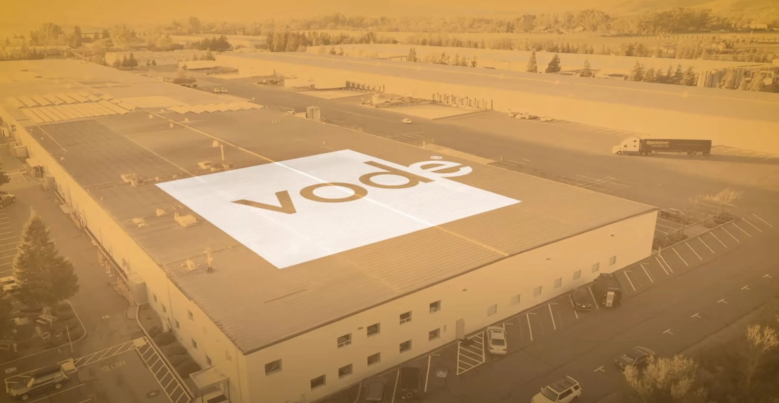 Aerial view of a large commercial building with a rooftop projection displaying the word "VOOK" during sunset, surrounded by a parking lot and open landscape.