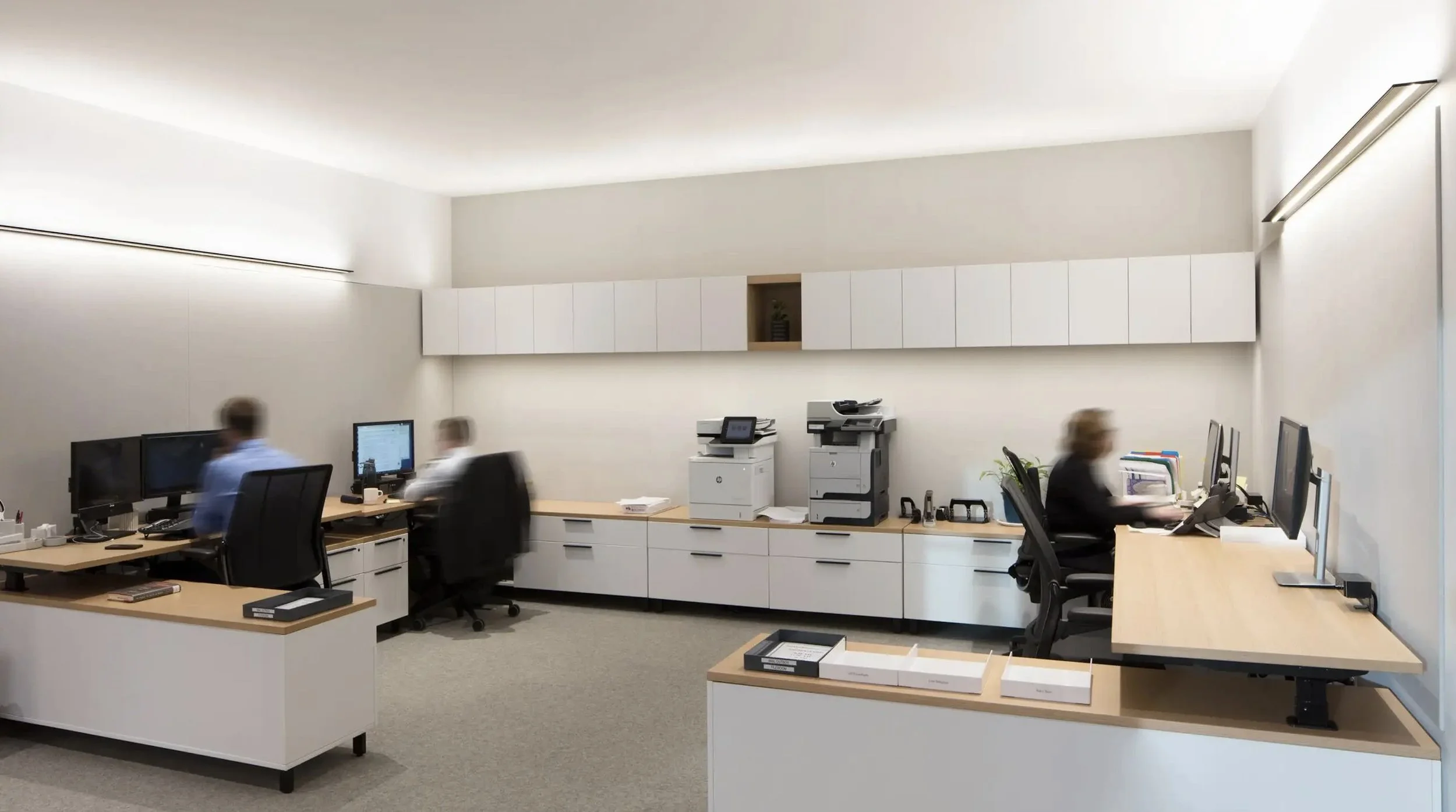 Office with three workers working at desks with multiple monitors, printers, and filing boxes, white walls, overhead storage cabinets, and minimal decor.
