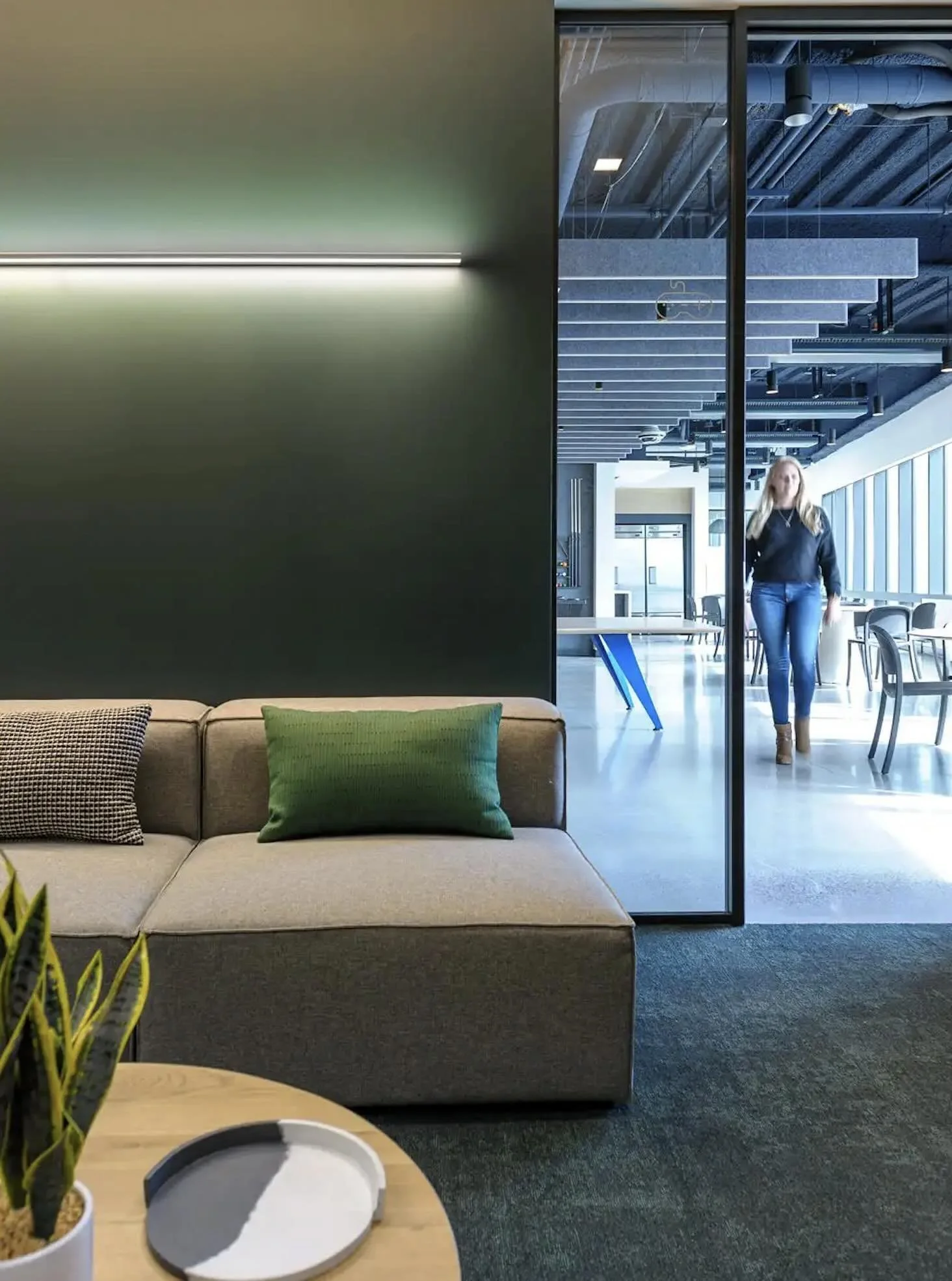 Modern office lounge area with a beige sofa and colorful pillows, a round wooden table with a plant and a decorative tray, and a glass wall revealing a woman walking in a bright, open workspace with large windows, chairs, and tables.