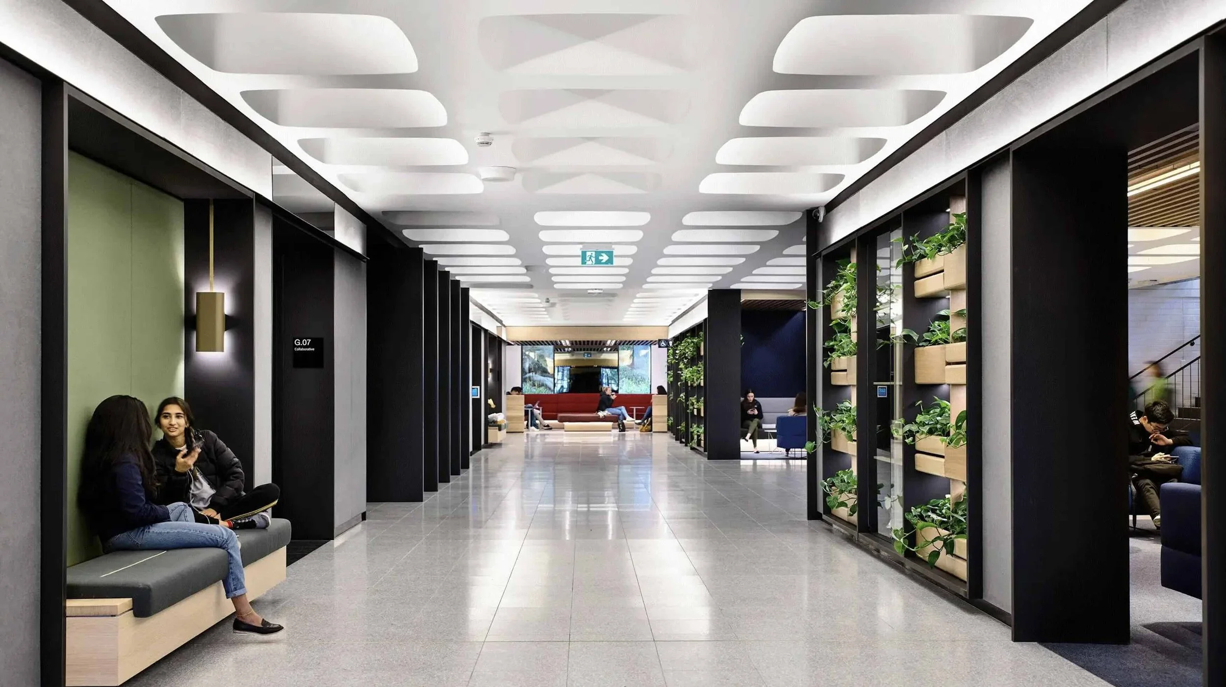 Modern airport or office lounge with seating areas, people seated and chatting or using their phones, decorative plants, and a well-lit ceiling.