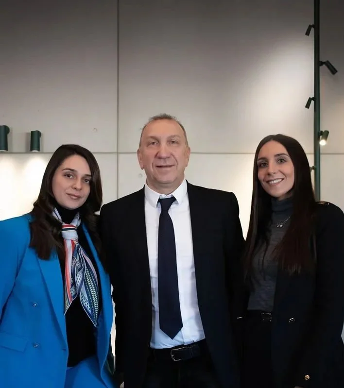 Three people, a man and two women, standing together indoors, smiling at the camera. The man is wearing a suit with a white shirt and dark tie, while the women are dressed in professional attire.