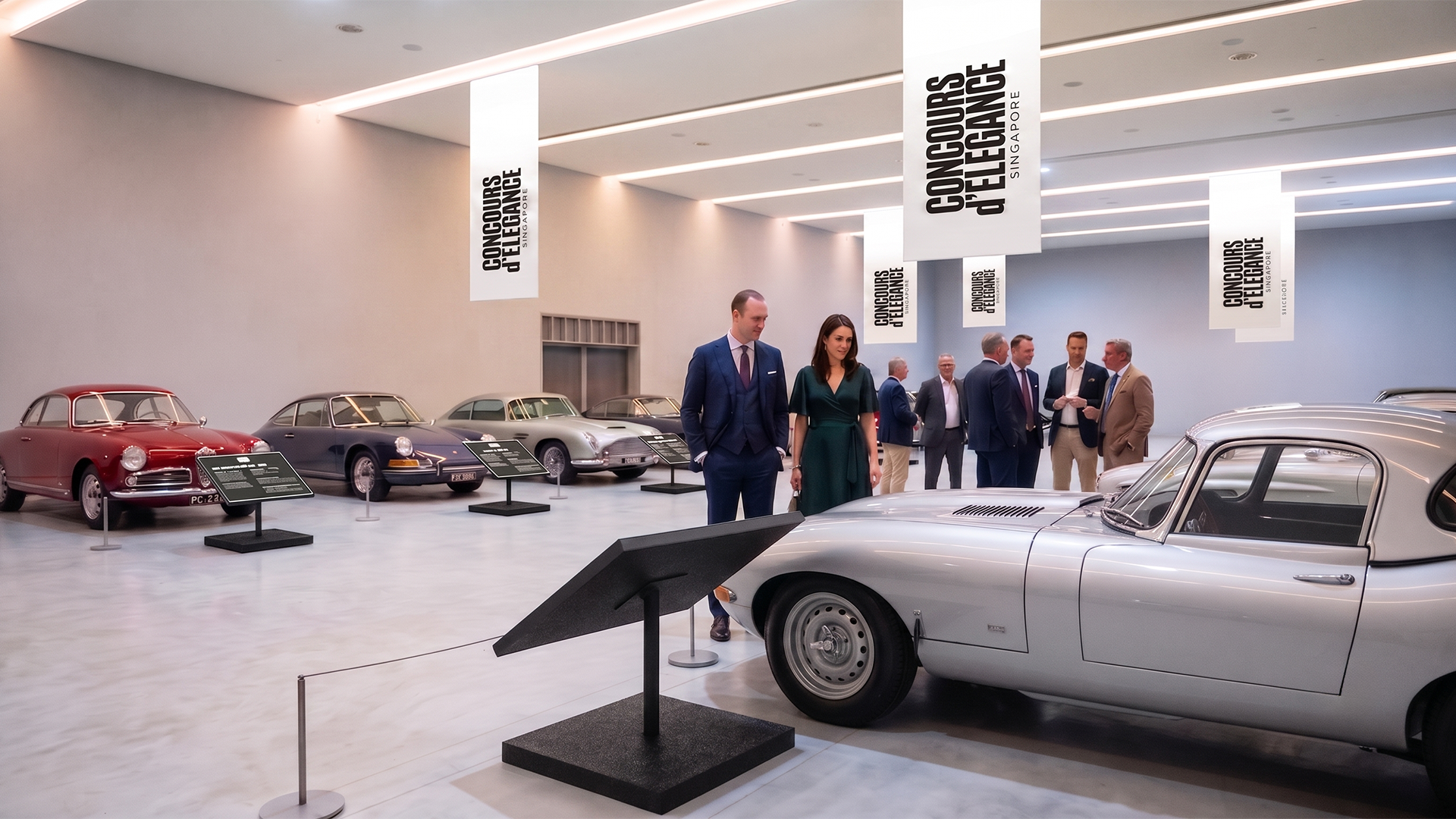 Classic cars on display at a car exhibition with people observing the vehicles and informational placards, held indoors with white walls and hanging banners.